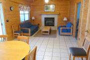 View of the living room and dining room of a modern cabin. There are two couches and a coffee table in the living room nestled in front of a fire place. The walls are wood and the floors are tile.