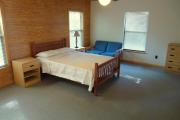 View of the bedroom in a modern cabin. There is a full size bed, a dresser, and a hide away couch that sleeps one person. The floors have short pile carpet and the walls are a combination of wood and white paint.