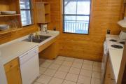 View of the kitchen of an ADA compliant modern cabin. The kitchen is equipped with accessible sink, stove, and dishwasher. The walls are wood and the floors are tile.
