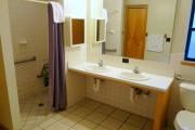 View of the bathroom in a modern cabin. There is an ADA compliant toilet, a dual sink vanity style counter with a large low mirror, and a walk-in/roll-in shower equipped with grab bars and a fold down chair. The walls are white and the floors are tile.