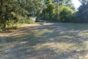 View of a vacant campsite with a picnic table, fire ring, and water spigot. The main area of the site is sandy and there are trees and bushes lining the back of the site. There is partial shade. This site does not have electricity.