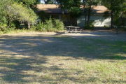 View of a vacant campsite with a picnic table, fire ring, and water spigot. The main area of the site is grassy with some sand and there are bushes on the edges of the site. There is partial shade. The campsite restroom is present in the background. This site does not have electricity.