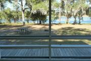 View from the porch of a historic cabin. A picnic table and fire ring is in the foreground and a line of trees and a beautiful blue lake are seen in the background.