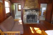 View of the inside of a historic cabin. There is one room with a bed, futon couch, and fire place. The floors and walls are wood and the fire place is made of stone. Just beyond the fireplace are the kitchen to the right and the bathroom to the left. These rooms are tiled and the walls are white.