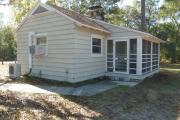 Close up of the front of an historic cabin. The cabin is grey with white trim and has a screen porch. There is a wooden boardwalk leading to the front porch. These cabins are wood frame constructed with lap siding.