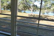 View from the porch of a historic cabin. A picnic table and fire ring is in the foreground and a line of trees and a beautiful blue lake are seen in the background.