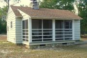 Close up of the front of an historic cabin. The cabin is grey with white trim and has a screen porch. There is a path leading to the front porch. These cabins are wood frame constructed with lap siding.