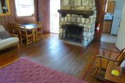 View inside an historic cabin. There is one room with a bed, futon, table, and fire place. The floors and walls are wood and the fire place is made of stone. Just beyond the fireplace are the kitchen to the right and the bathroom to the left. These rooms are tiled and the walls are white