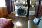 View inside an historic cabin. There is one room with a bed, futon, dresser, and fire place. The floors and walls are wood and the fire place is made of stone. Just beyond the fireplace are the kitchen to the right and the bathroom to the left. These rooms are tiled and the walls are white.