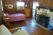 View inside an historic cabin. There is one room with a bed, dresser, futon, and fire place. The floors and walls are wood and the fire place is made of stone. Just beyond the fireplace is the bathroom. The bathroom is tiled and the walls are white.