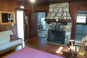 View inside an historic cabin. There is one room with a bed, dresser, futon, and fire place. The floors and walls are wood and the fire place is made of stone. Just beyond the fireplace are the kitchen to the left and the bathroom to the right. These rooms are tiled and the walls are white.