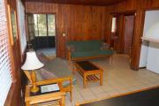 View of the inside of a block cabin. The living room has two futons, a coffee table, and a dining table. Just beyond the living room are the bedroom and bathroom. Also pictured is the screen porch off of the living room. The walls are wood and the floors are tile.