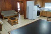 View of the inside of a block cabin. The living room has two futons, a coffee table, and a dining table. Just beyond the living room are the bedroom and bathroom. Also pictured is the kitchen. The walls are wood and the floors are tile.
