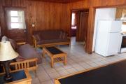View of the inside of a block cabin. The living room has two futons, a coffee table, and a dining table. Just beyond the living room are the bedroom and bathroom. Also pictured is the kitchen. The walls are wood and the floors are tile.