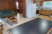 View of the inside of a block cabin. The living room has two futons, a coffee table, and a dining table. Just beyond the living room are the bedroom and bathroom. Also pictured is the kitchen. The walls are wood and the floors are tile.