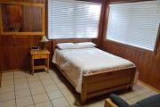View of the bedroom in a block cabin. The bedroom has a full size bed and a nightstand. The floors are tile and the walls are wood. Two large picture windows create wonderful amounts of natural light for the room.
