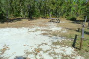 View of a vacant site with a picnic table and fire ring. There are two wooden barricades on the left edge of the site. The main part of the site is sandy with sparse vegetation. There is a mix of trees in the background to include long leaf pine and turkey oak. The site is in full sun.