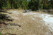View of a vacant site with picnic table, fire ring, electric pedestal, and water spigot. There is a wooden barricade on the left edge of the site. The main part of the site is sandy with sparse vegetation. There is a mix of trees in the background. The site is in full sun.
