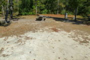 View of a vacant campsite with a fire ring, a picnic table, electric pedestal, and a water spigot. The main part of the site is sand. Grass and a small tree are located on the right edge of the site and there are two medium sized trees on the left edge. The site has partial shade.
