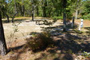 View of a vacant campsite with a fire ring, a picnic table, electric pedestal, and a water spigot. The vegetation is sparse and there are several trees on the edges of the site. A few small bushes are present in the foreground and there is partial shade in the site.