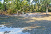 View of a vacant campsite with a fire ring, picnic table, electric pedestal, and a water spigot. The main part of the site is grassy and well shaded. There are several trees on the back edge of the site.