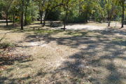 View of a vacant site showing a brown site marker with an arrow on a wooden post. The site is grassy with some patches of sand. There is a fire ring, picnic table, electric pedestal, and a water spigot. The site has partial shade and a few small trees in the center of the site.