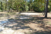 View of a vacant site showing a brown site marker with an arrow on a wooden post. The site is grassy with some patches of sand. There is a fire ring, picnic table, electric pedestal, and a water spigot. The site has partial shade and a few small trees in the center of the site.
