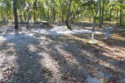 View of a vacant site with a picnic table, fire ring, electric pedestal, and a water spigot. The main part of the site is sandy with sparse vegetation. There are a few medium size trees in the center of the site and the site is well shaded.