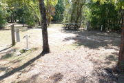 View of a vacant site with a picnic table, fire ring, electric pedestal, and a water spigot. Grass is present in most of the site and there is partial shade. The site has several small trees on the edges.