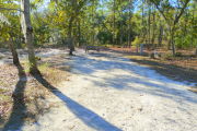 View of a vacant campsite with a picnic table, fire ring, electric pedestal, and a water spigot. The main part of the site is sandy with medium size trees on both sides. There are multiple trees at the back and the site is well shaded.