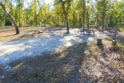View of a vacant campsite with a picnic table, fire ring, electric pedestal, and a water spigot. The main part of the site is sandy with medium size trees on both sides. There are multiple trees at the back and the site is well shaded.