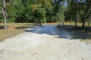 View of a vacant site with a picnic table, fire ring, electric pedestal, and a water spigot. The majority of the site is sandy with a grassy area toward the back. There are a few trees on the left and right edges and the site is partially shaded. The back edge of the site is lined with trees.