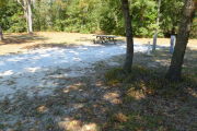 View of a vacant site with a picnic table, fire ring, electric pedestal, and a water spigot. The majority of the site is sandy with a grassy area toward the back. There are a few trees on the left and right edges and the site is partially shaded. The back edge of the site is lined with trees.