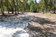 View of a vacant site with a picnic table, fire ring, electric pedestal, and a water spigot. The site is sandy with sparse vegetation. There is a group of six trees to the left of the site and the back of the site is lined with trees. There is partial shade in the site.