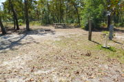 View of a vacant site with a picnic table, fire ring, electric pedestal, and a water spigot. The site is sandy with sparse vegetation. There is a group of six trees to the left of the site and the back of the site is lined with trees. There is partial shade in the site.