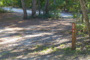View of a vacant site with a brown site marker with an arrow on a wooden post. The main part of the site is sandy with sparse vegetation. The site marker is in the foreground and there is a group of five large trees in the background.