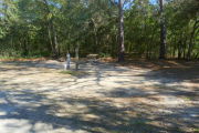 View of a vacant site with a picnic table, fire ring, electric pedestal, and a water spigot. The site is sandy with sparse vegetation. A variety of trees are on the edges of the site and the site is shaded.