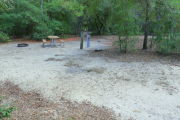 View of a vacant campsite with a picnic table, fire ring, electric pedestal, and water spigot. The main part of the site is sandy. There are several trees surrounding the site and it is partly shaded.