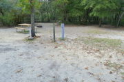 View of a vacant campsite with a picnic table, fire ring, electric pedestal, and water spigot. The main part of the site is sandy. There are several trees surrounding the site and it is partly shaded. A medium size tree is located in the middle of the site.