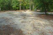 View of a vacant campsite with a picnic table, fire ring, electric pedestal, and water spigot. The site is sandy and has trees all around. There is partial shade.
