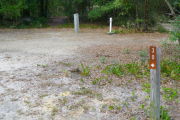 Close up of a brown site marker with an arrow on a wooden post. The foreground is sandy with some vegetation. An electric pedestal and water spigot are in the background and there is dense vegetation beyond the back edge of the site.