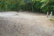 View of a vacant campsite with a picnic table, fire ring, electric pedestal, and water spigot. The site is sandy and has trees all around. There is partial shade.