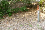 Close up of a brown site marker with an arrow on a wooden post. The foreground is sandy and there are some small trees in the background.