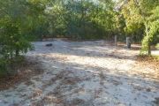 View of a vacant campsite with a fire ring and electric pedestal. The main part of the site is sandy with some grass around the edges. The site is lined with trees and the back of the site is densely forested.