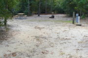 View of a vacant campsite with a picnic table, fire ring, water spigot, and electric pedestal. The main part of the site is sandy with some grass around the edges. A few small trees are present within the site. The site is lined with trees and the back is densely forested.