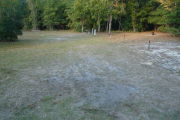 View of a vacant campsite with a picnic table, fire ring, electric pedestal, and a water spigot. The site is grassy with a few sandy areas. Three wooden posts with red reflective tape are on the right edge of the site and a wooden barricade at the back. The back of the site is densely forested.
