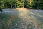 View of a vacant campsite with a picnic table, fire ring, electric pedestal, and a water spigot. The site is grassy with a few sandy areas. Three wooden posts with red reflective tape are on the right edge of the site and the back of the site is densely forested. There is partial shade.