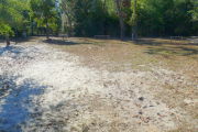 View of a vacant campsite with a picnic table, fire ring, electric pedestal and water spigot. Several wooden barricades are on the edges of the site. The main part of the site is grassy with some patches of sand. The back of the site is densely forested and there is partial shade.