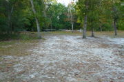 View of a vacant campsite with a picnic table, fire ring, water spigot, and electric pedestal. The site is sandy with large areas of grass in the back and on the sides. There are two trees on each side of the entrance. Wooden barricades are present on the edges and the site is partially shaded.
