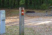 Close up of a brown site marker on a wooden post. There is an electric pedestal to the left of the marker and there is a fire ring and picnic table in the background.